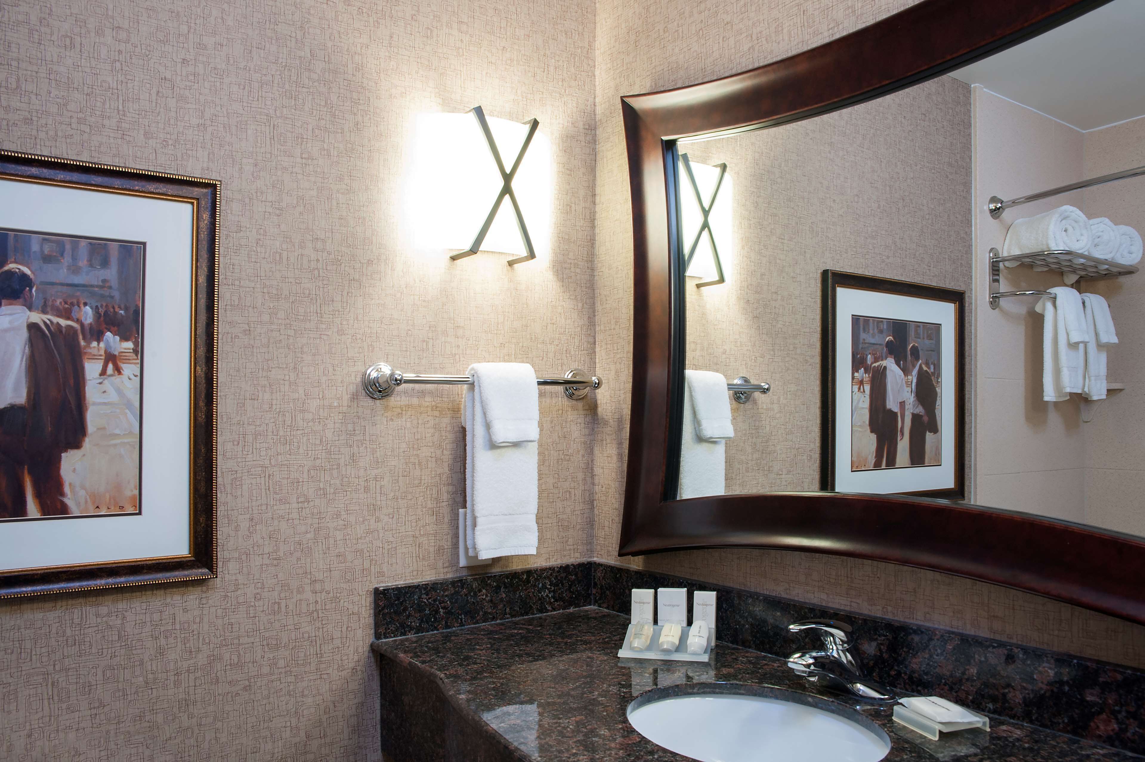 A hotel bathroom with a large mirror, framed artwork, a towel rack, and a sink with toiletries.