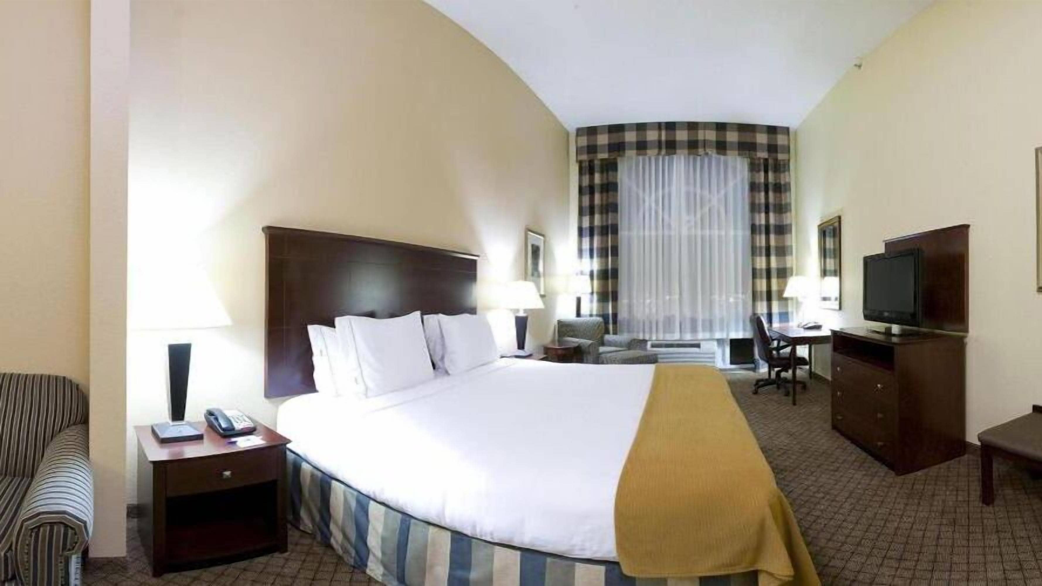 Una habitación de hotel con una cama grande, un escritorio con silla, televisión y ventana con cortinas.
