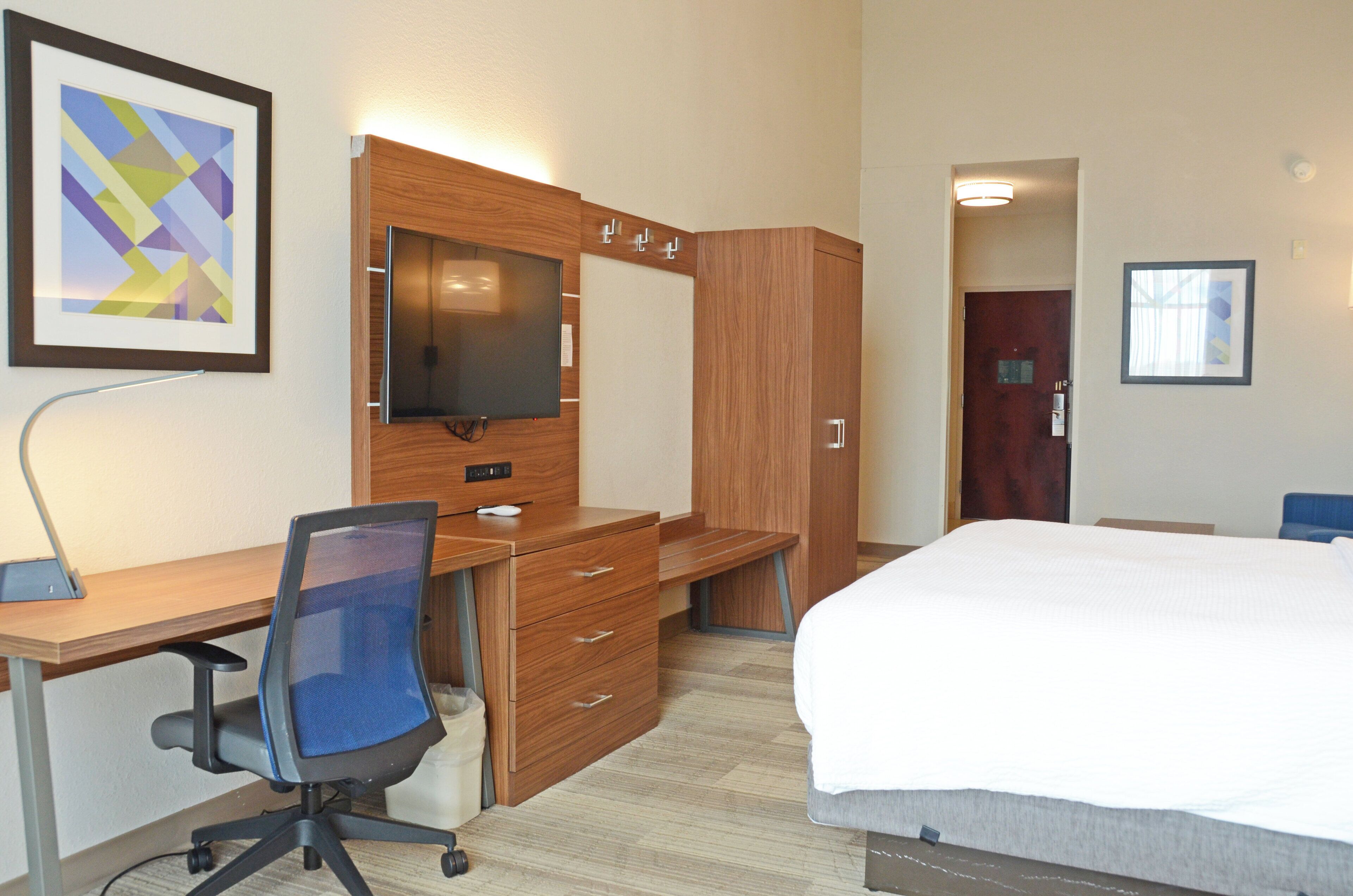 A hotel room with a wooden desk, a chair, a bed, a TV, and a wardrobe.