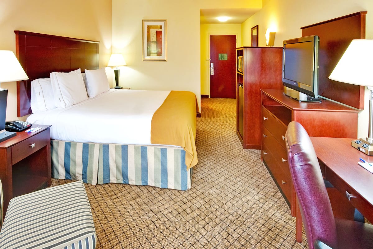 A hotel room with a bed, bedside table, lamp, TV, desk, chair, and a mirror on the wall.