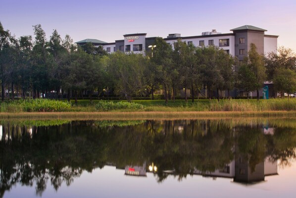 A Courtyard hotel with a reflection in a calm body of water, surrounded by trees and greenery. - Courtyard by Marriott Miami at Dolphin Mall (Miami)