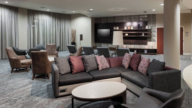 Eine moderne Hotelhalle mit Ecksofa, mehreren Sesseln, einem runden Couchtisch und einem an der Wand befestigten Flachbildfernseher.