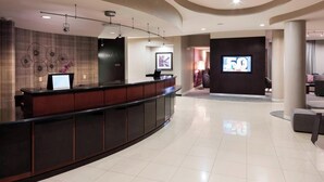 Eine moderne Hotellobby mit einer Rezeption, einem Bildschirm, der '5G' anzeigt, und einer Sitzecke mit einer Couch und Sesseln.