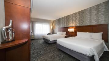 A hotel room with two beds, a wooden headboard, a nightstand, a lamp, and a large window with curtains.