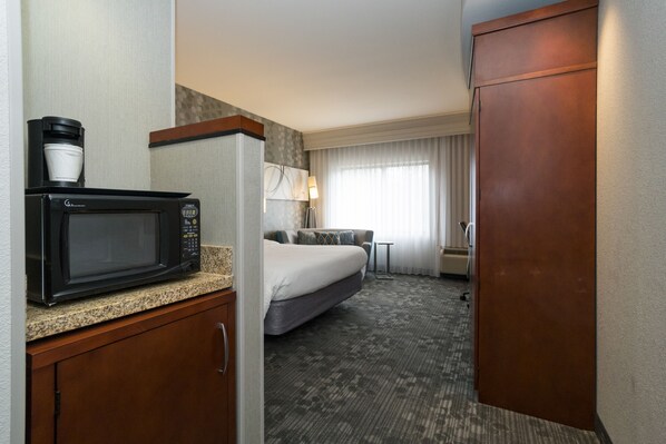 A hotel room with a kitchenette area including a microwave, coffee maker, and a bed with a gray mattress and pillows.