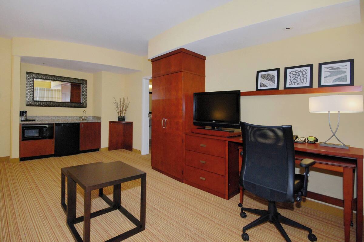 A compact hotel room with a kitchenette, a wooden desk, a chair, a television, and a small table.