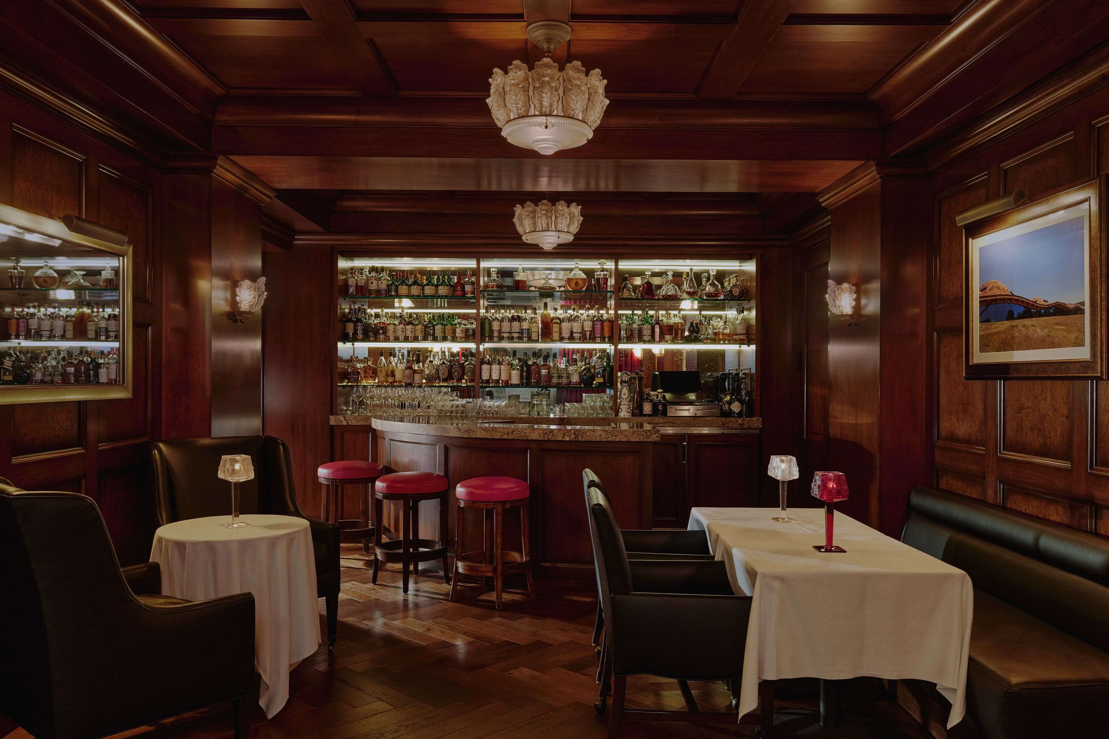 A bar area with wooden paneling, a chandelier, and a well-stocked bar. There are tables and stools for seating.