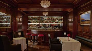 A bar area with wooden paneling, a chandelier, and a well-stocked bar. There are tables and stools for seating.