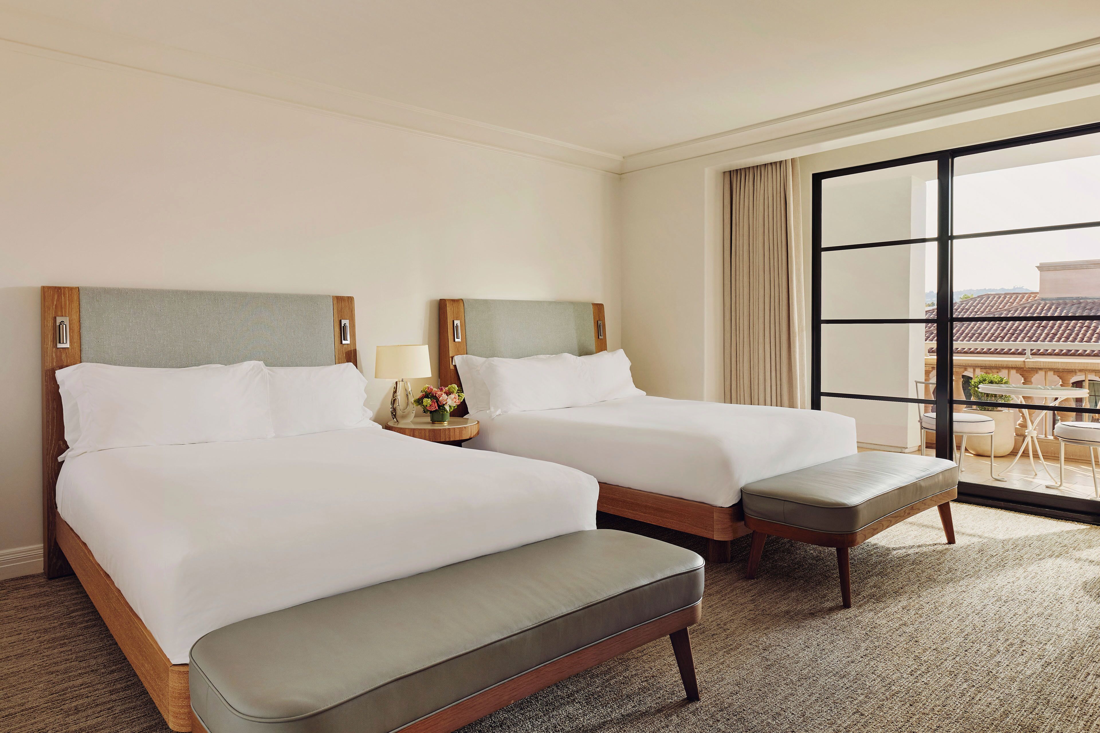 A hotel room with two beds, a bench, a nightstand with a lamp, and a large window with a view of the outside.