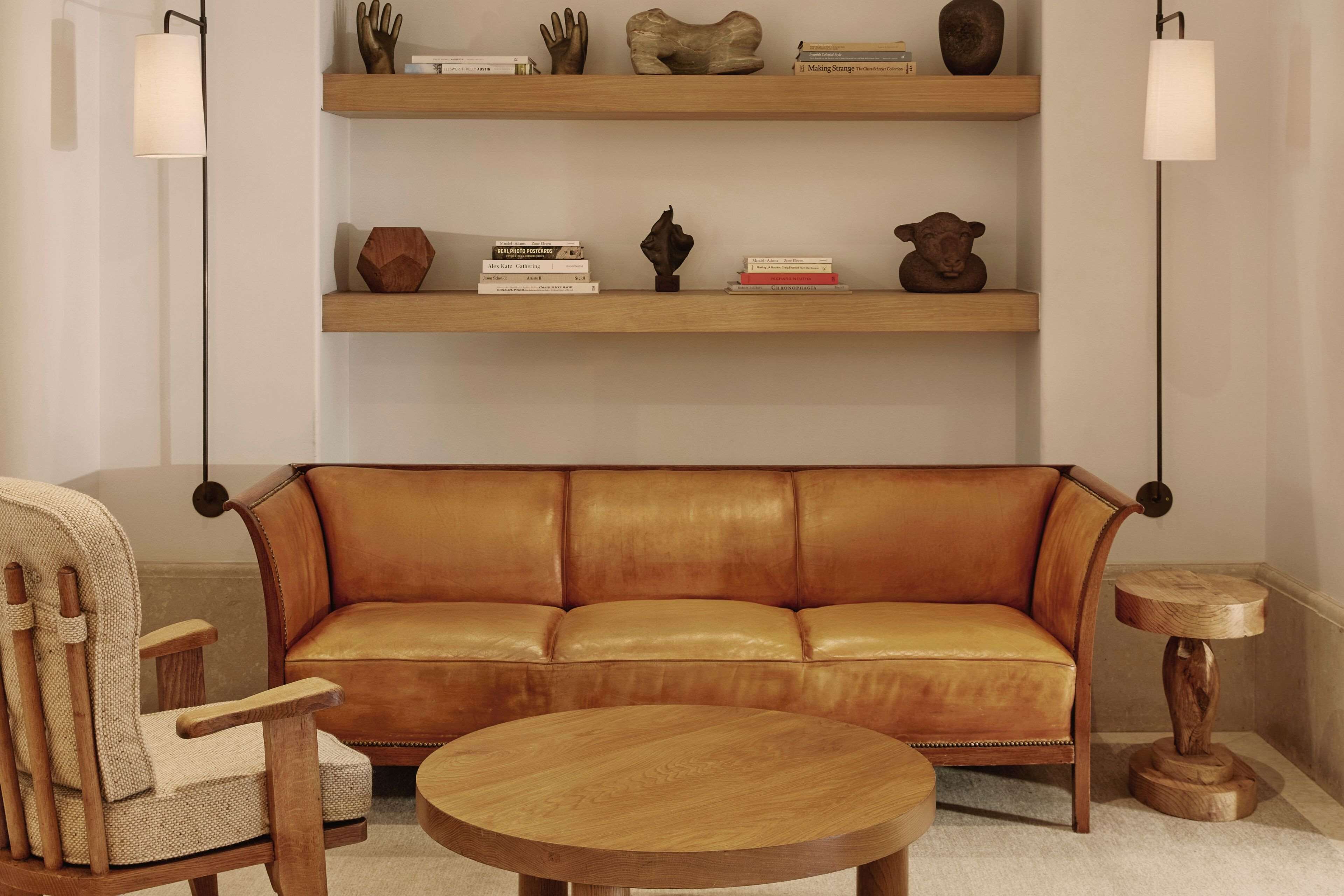 A living room with a leather sofa, wooden side table, and a round coffee table. There are two shelves above the sofa with decorative items and books.