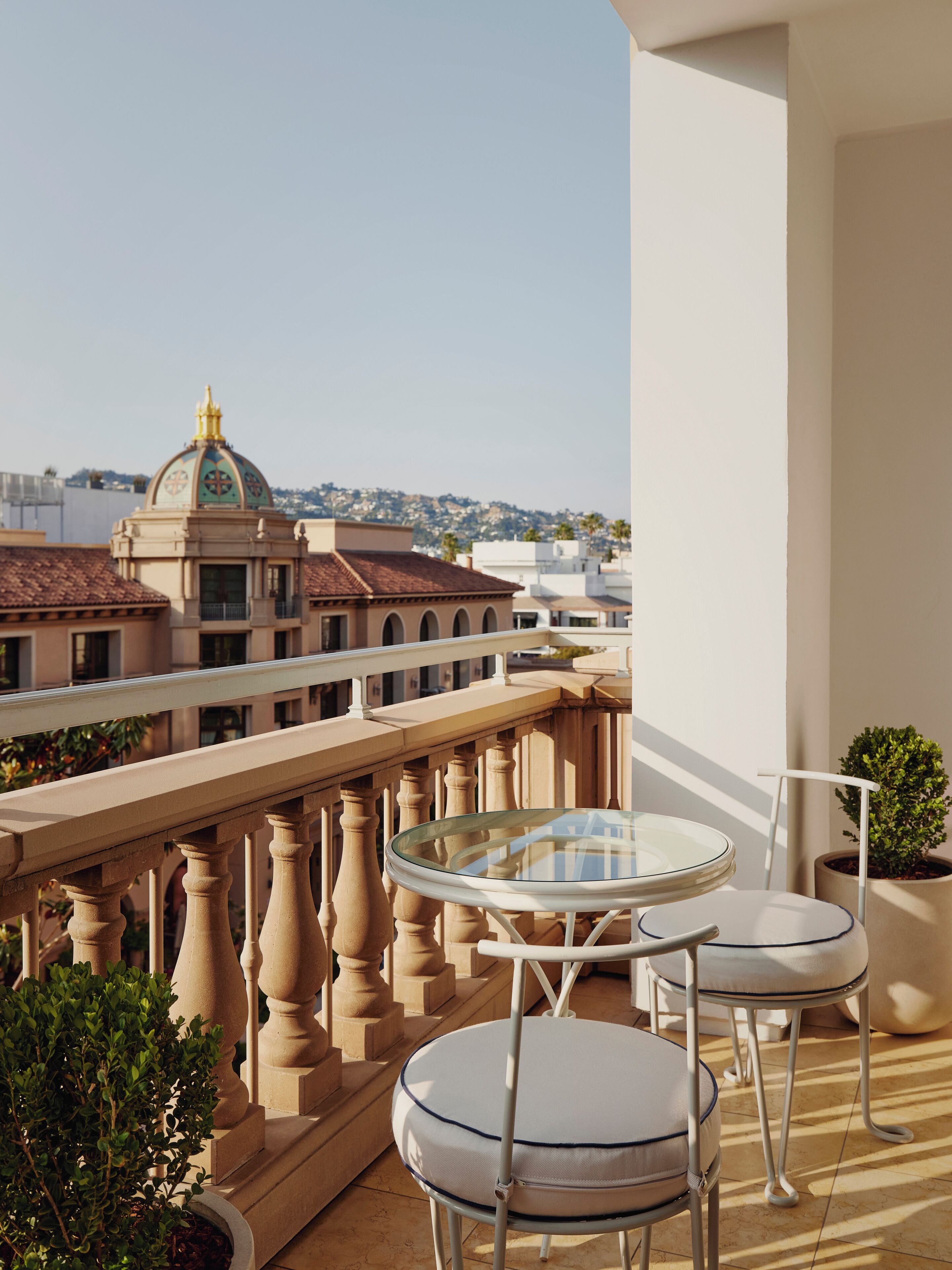 A balcony with a glass table and two chairs, overlooking a cityscape with historic buildings and a hill in the distance.