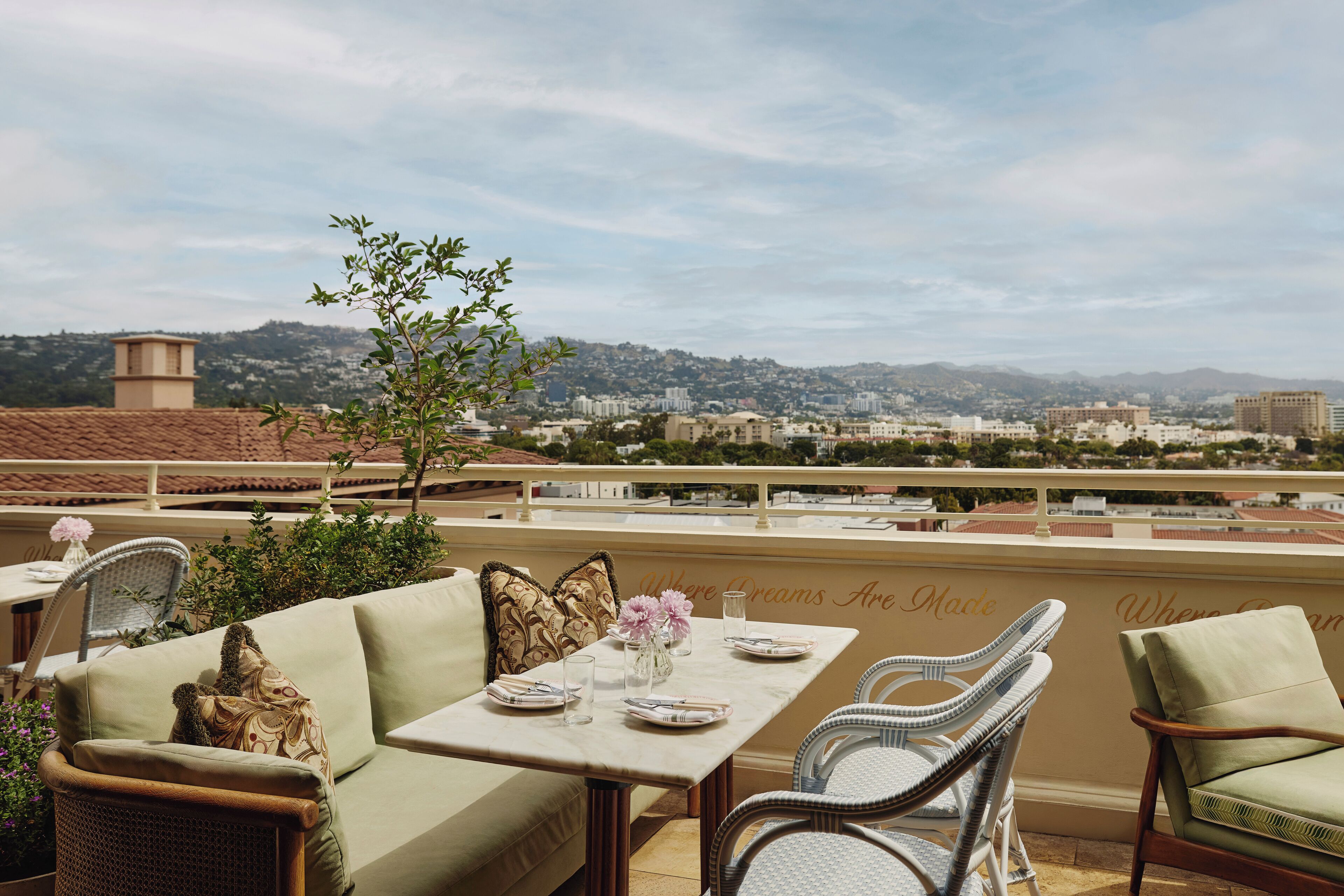 Une terrasse avec une table dressée pour le dîner, des chaises en osier, un canapé confortable et une vue sur un paysage urbain avec des bâtiments et des collines en arrière-plan.