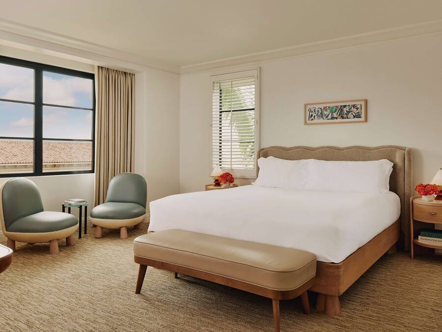 A hotel room with a large bed, two chairs, a bench, a small table, a nightstand, and a window with curtains.