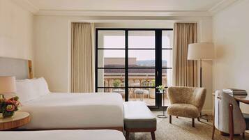 A hotel room with two beds, a chair, a desk, and a view of the city through large windows.