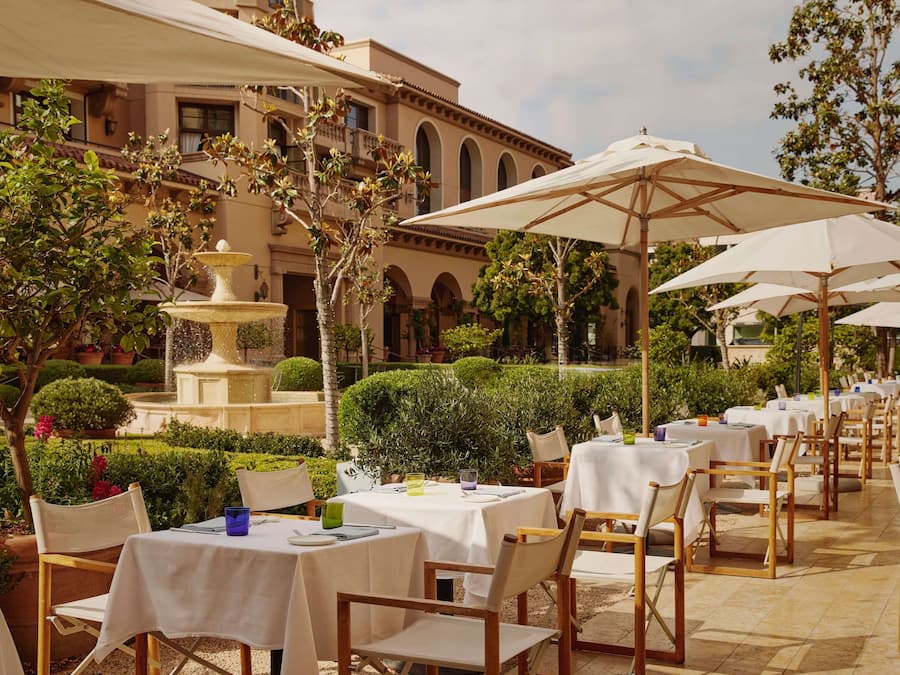 Un área de comedor al aire libre con mesas y sillas, una fuente y un edificio con arcos al fondo.