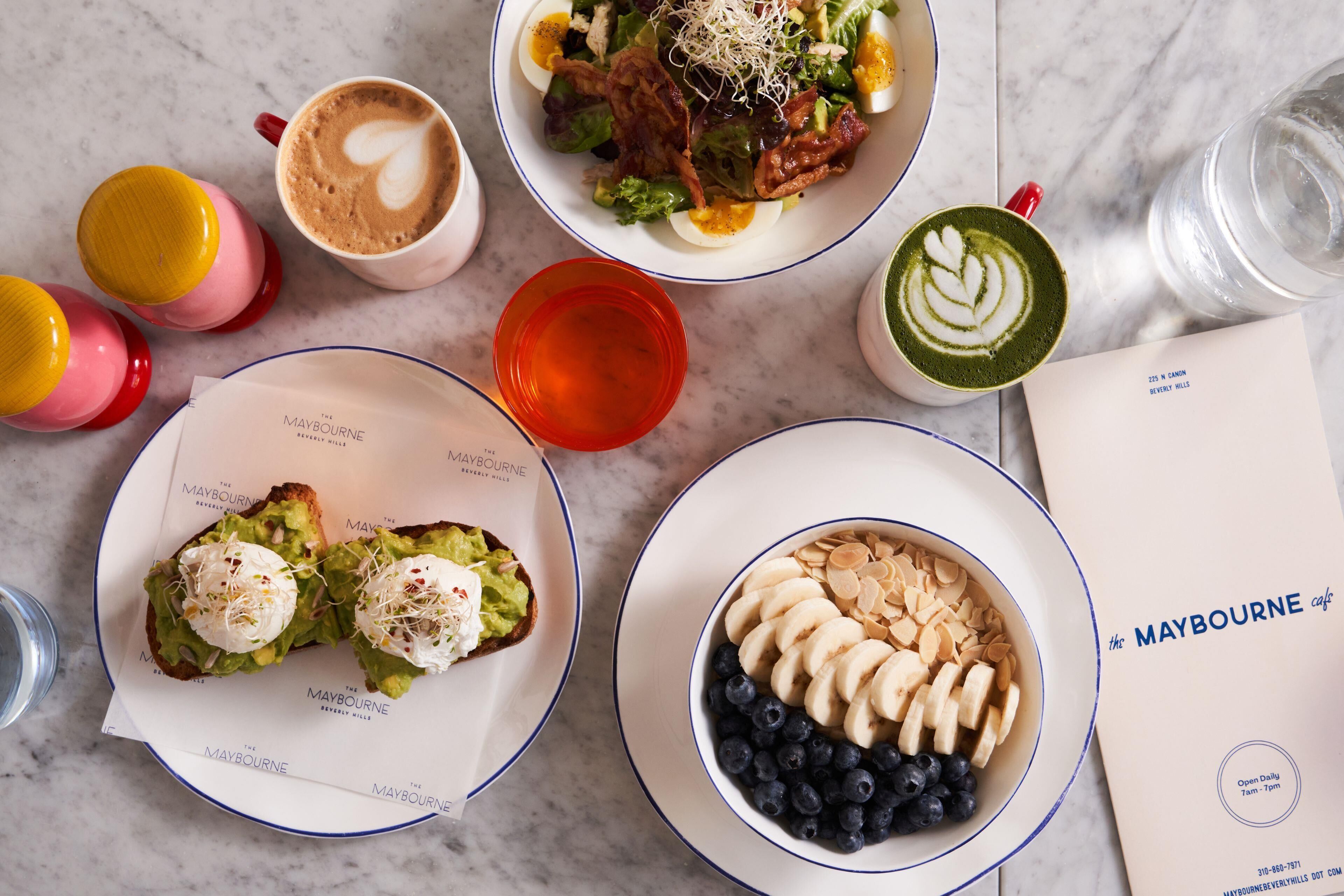 Una mesa con diversos alimentos y bebidas, como una ensalada, tostadas de aguacate, un tazón de fruta y tazas de café.