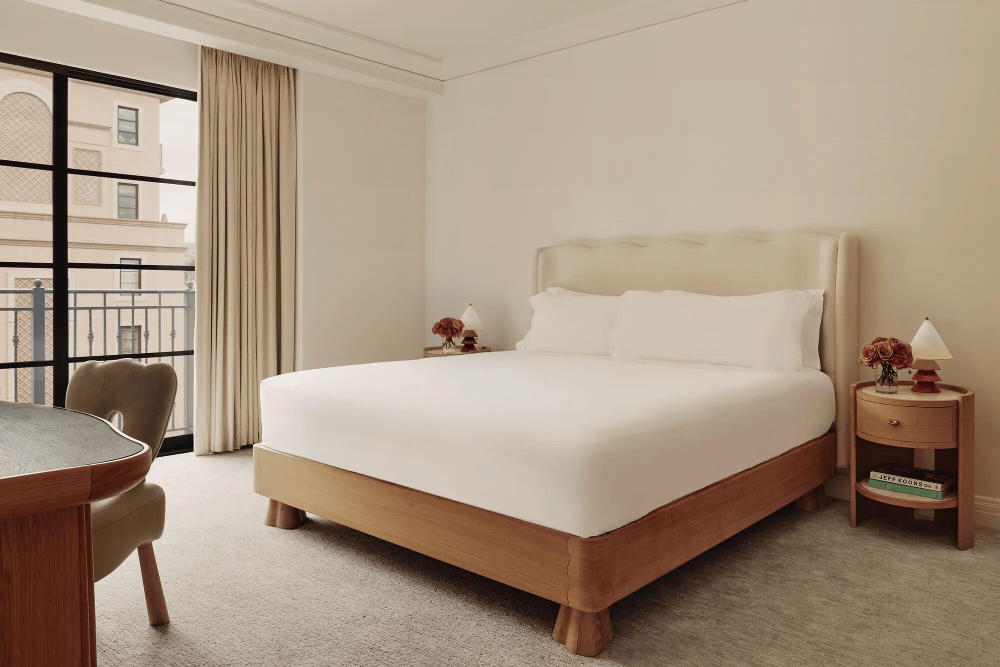 Ein Hotelzimmer mit einem großen Bett, einem Schreibtisch und einem Stuhl. Es gibt ein Fenster mit Vorhängen, einen Nachttisch mit einer Lampe und einen Blick auf Gebäude.