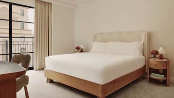 Ein Hotelzimmer mit einem großen Bett, einem Schreibtisch und einem Stuhl. Es gibt ein Fenster mit Vorhängen, einen Nachttisch mit einer Lampe und einen Blick auf Gebäude.