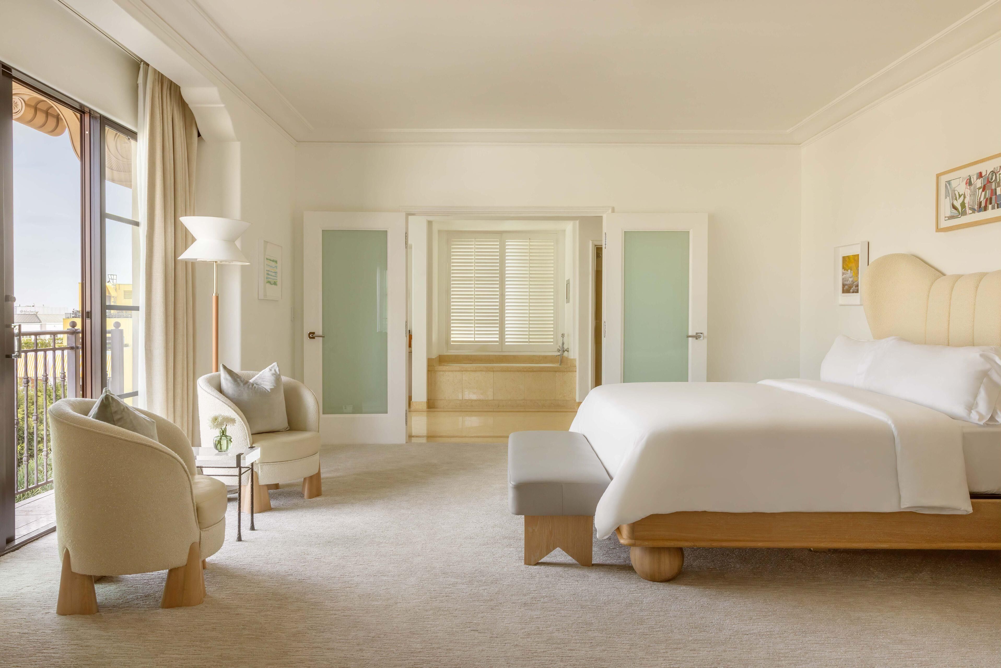 Habitación de hotel con una cama grande, zona de estar con una silla y una mesa, vistas al exterior a través de una ventana y una puerta que da a un baño.