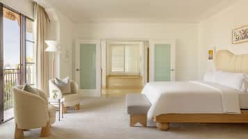 Habitación de hotel con una cama grande, zona de estar con una silla y una mesa, vistas al exterior a través de una ventana y una puerta que da a un baño.