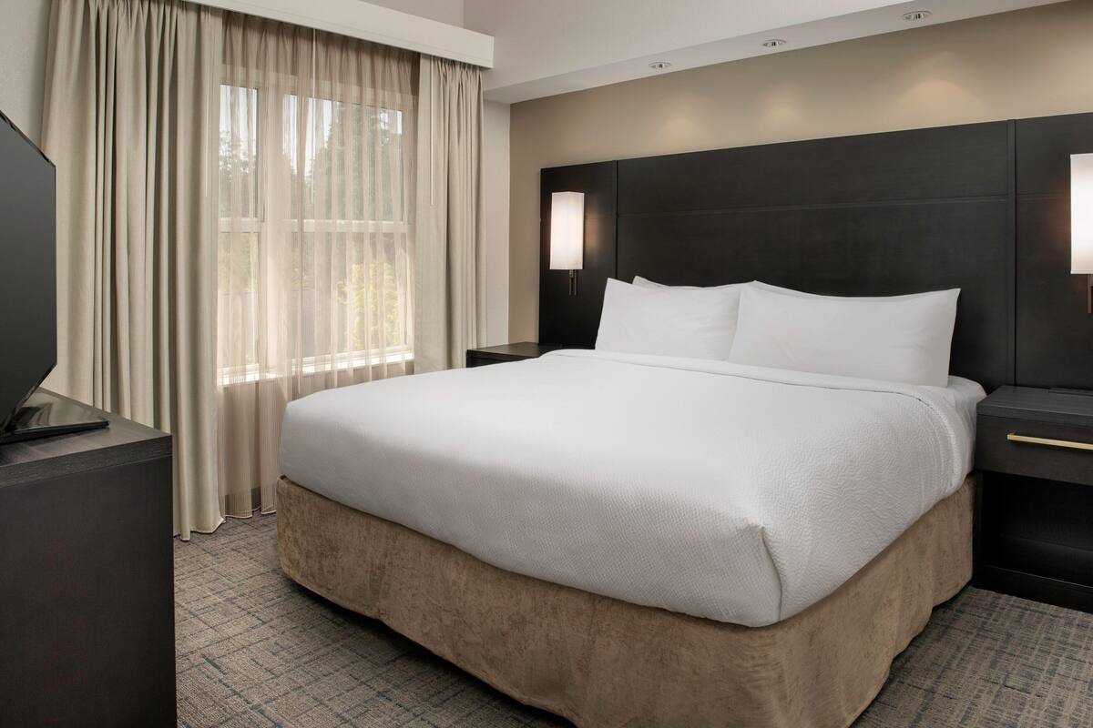 Une chambre d’hôtel avec un grand lit, un téléviseur posé sur un meuble, une table de chevet et une fenêtre avec des rideaux.