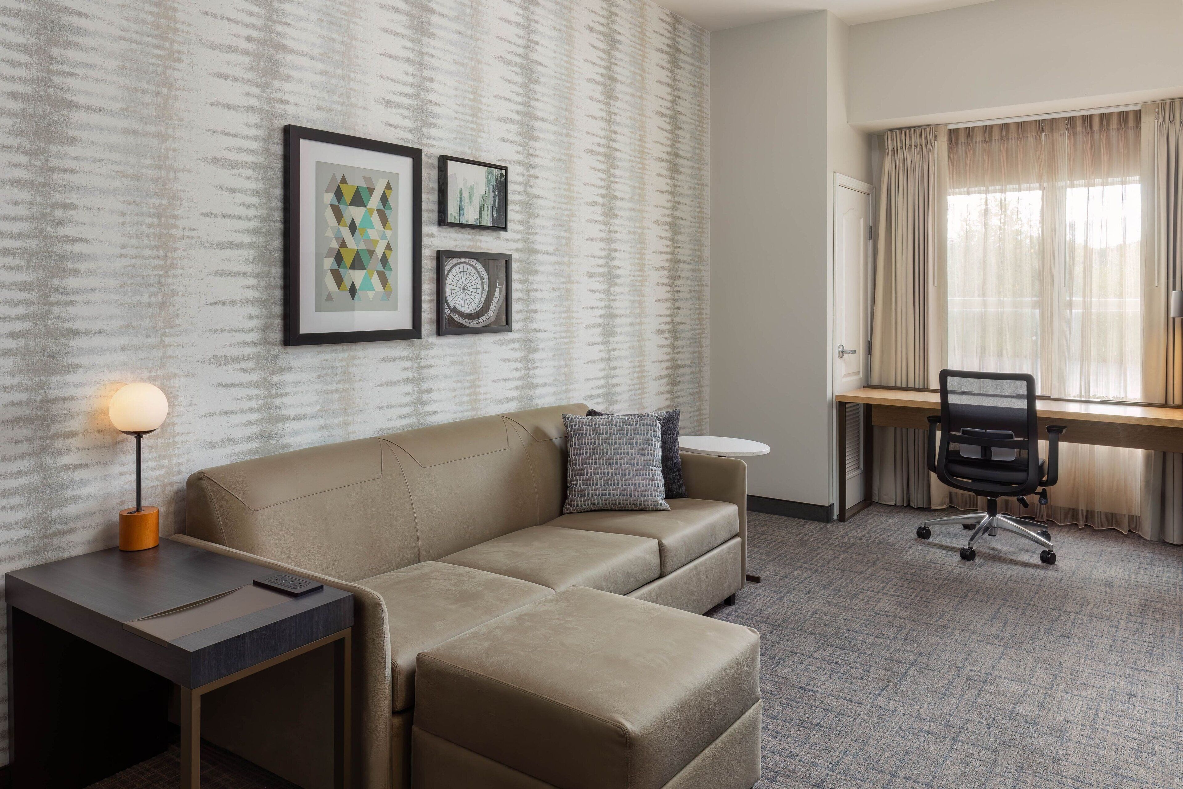 Habitación de hotel con sofá, una mesita con lámpara, escritorio y silla, y cuadros en la pared.