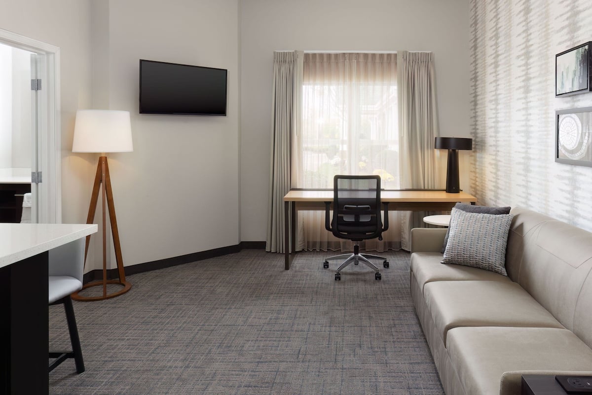 A hotel room with a desk, chair, sofa, and a TV mounted on the wall.