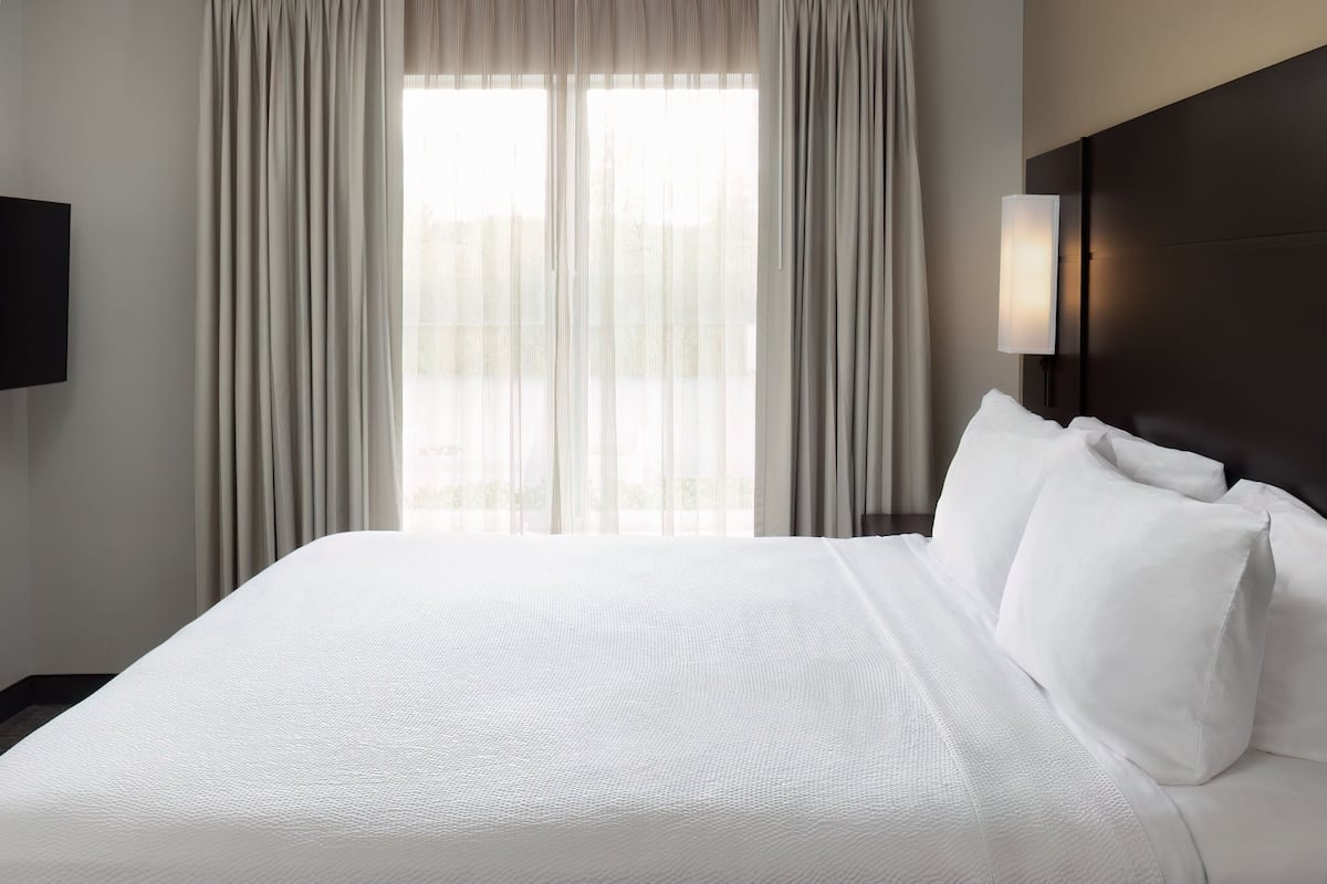 A hotel room with a large bed, a flat-screen TV, and a window with sheer curtains.