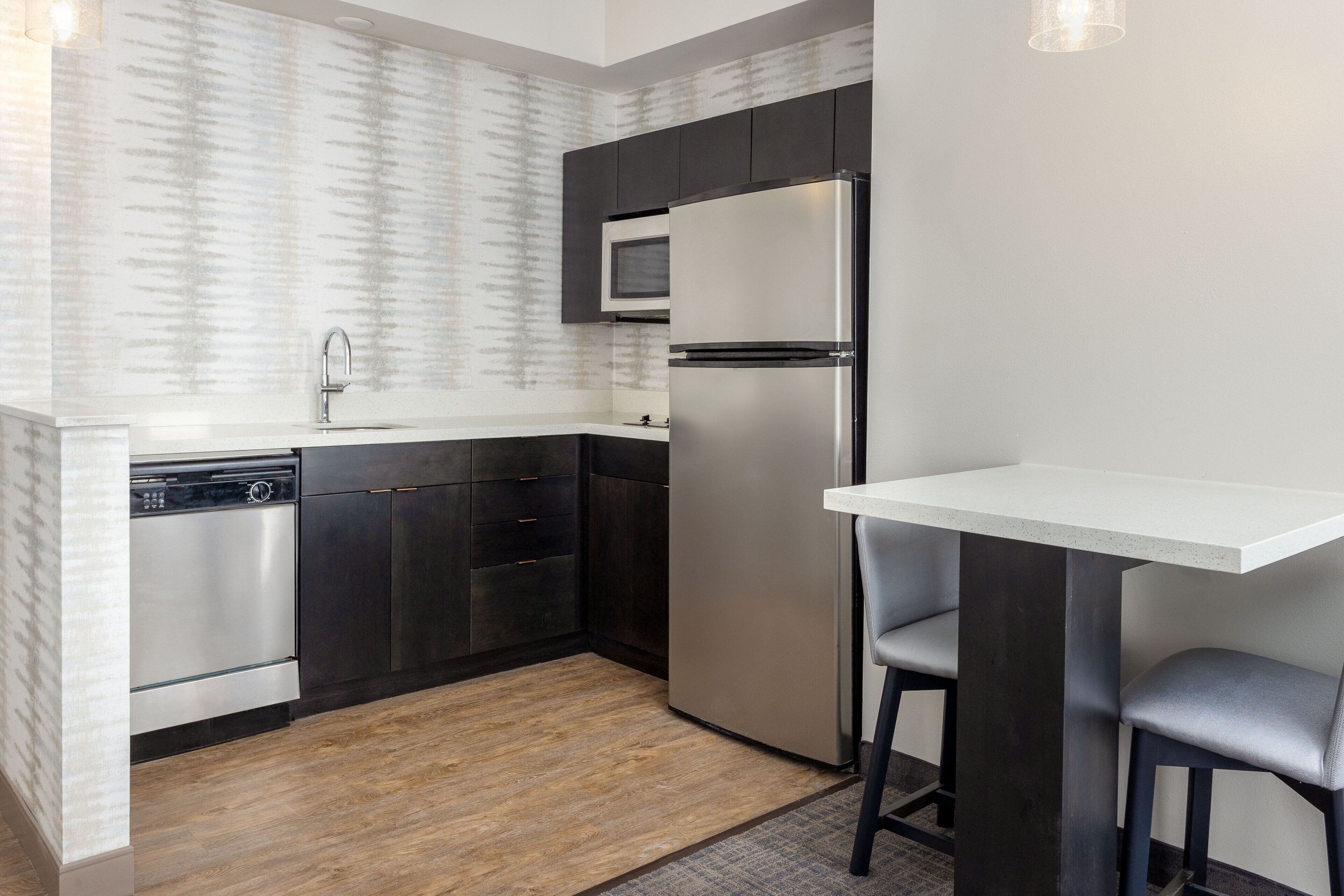A modern kitchen with dark cabinetry, a stainless steel refrigerator, a built-in microwave, and a dishwasher. There is a small bar area with a white countertop and two bar stools.