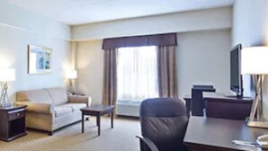 Habitación de hotel con cama, sofá, escritorio y silla. Se ve una ventana con cortinas y una lámpara sobre el escritorio.