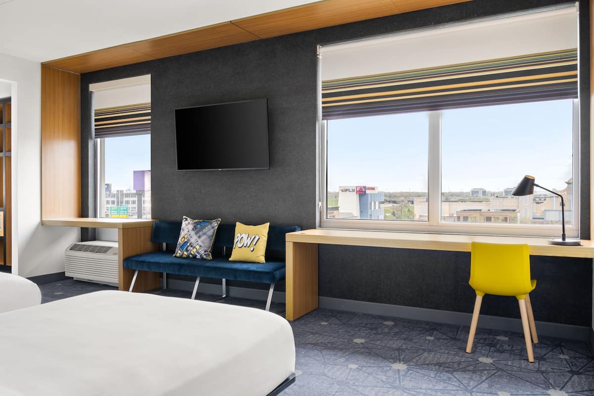 Quarto de hotel moderno com cama, escrivaninha com cadeira, sofá com almofadas, televisão de tela plana e janelas amplas com vista para a cidade.
