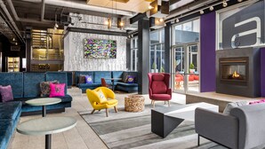 A modern lounge area with sofas, a fireplace, and a large window offering a view of the outdoors. - Aloft by Marriott Chicago O'Hare (Rosemont)