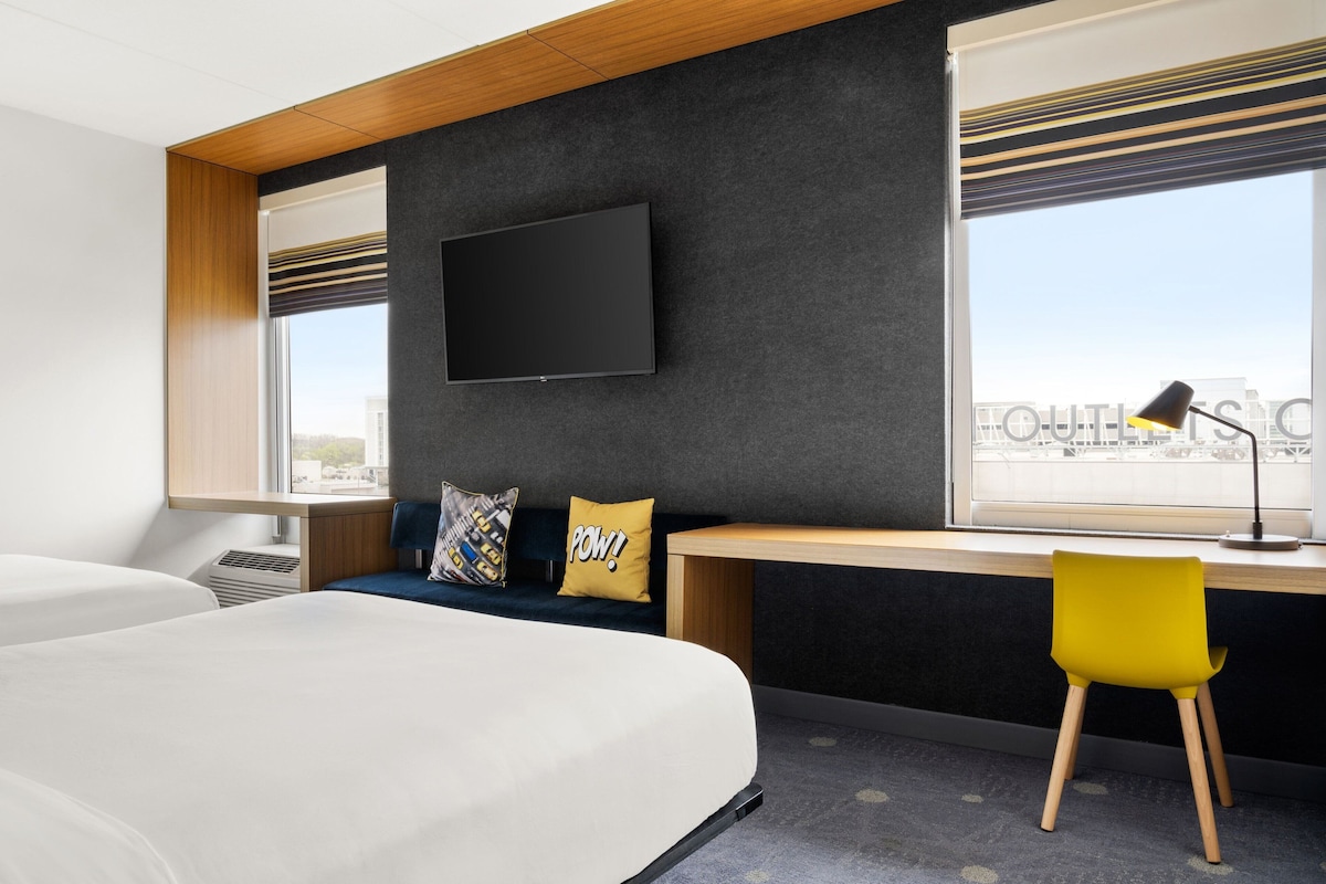 A modern hotel room with two beds, a desk, a chair, a TV, and large windows.