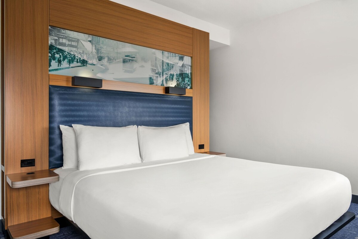 A modern hotel room with a large bed, wooden headboard, and a cityscape artwork above the bed.