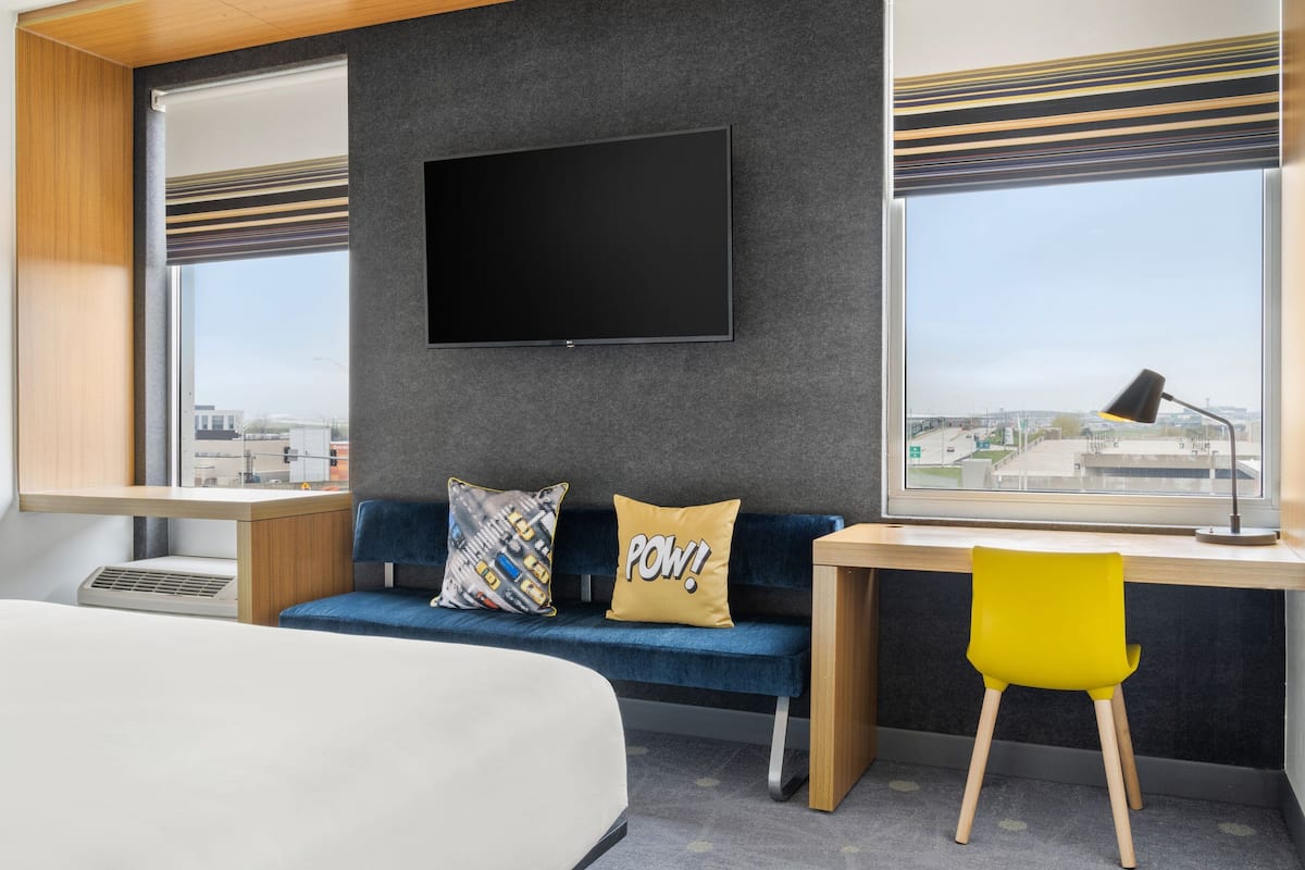 Quarto de hotel moderno com uma cama, escrivaninha, sofá azul e televisão fixada na parede.