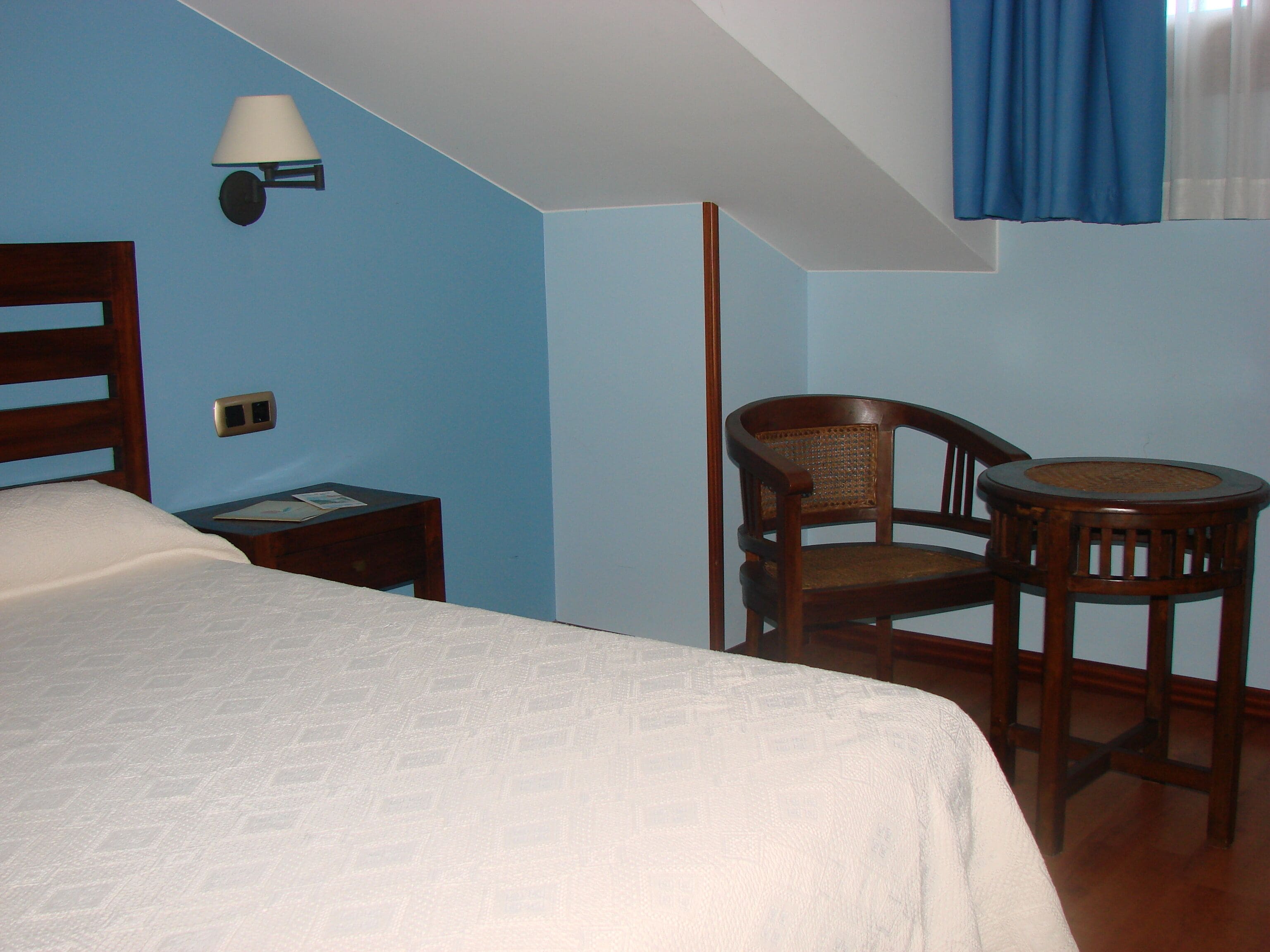Una habitación con una cama, una silla, una mesita, una lámpara y una ventana con cortinas.