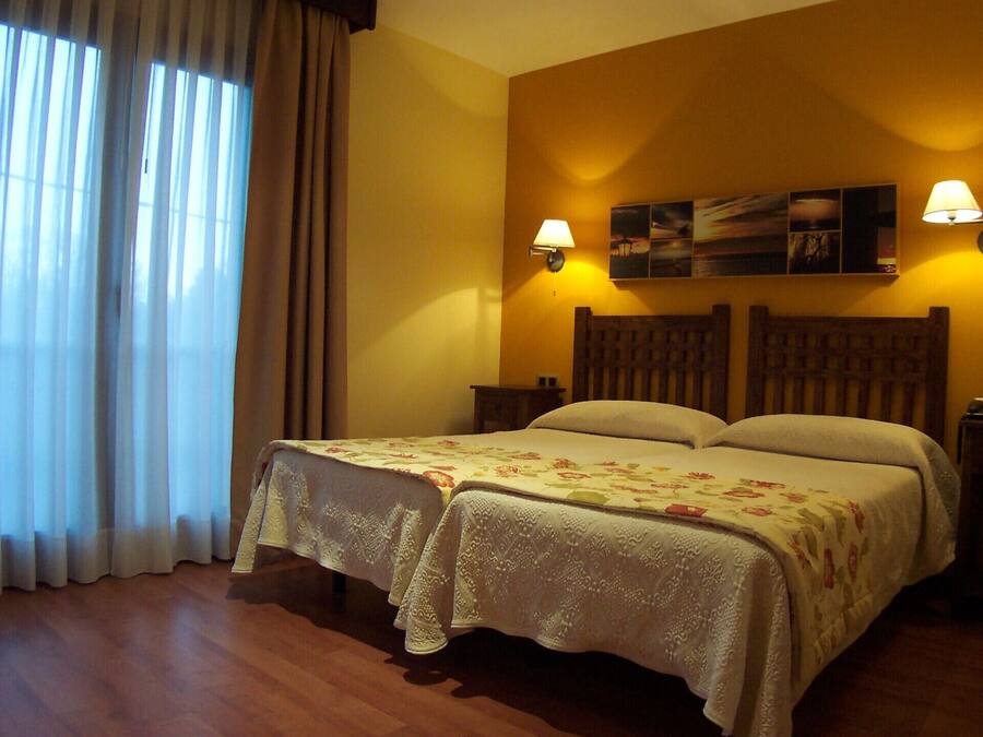Una habitación de hotel con una cama, dos mesitas de noche, una lámpara, un cuadro en la pared y una ventana con cortinas.