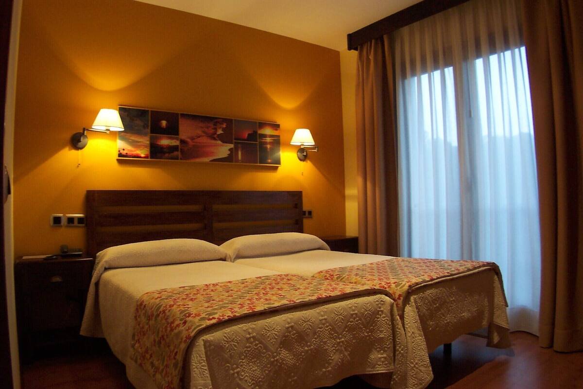 Une chambre d’hôtel avec un lit double, deux lampes fixées au mur, une table de chevet, une fenêtre avec des rideaux et un tableau décoratif.