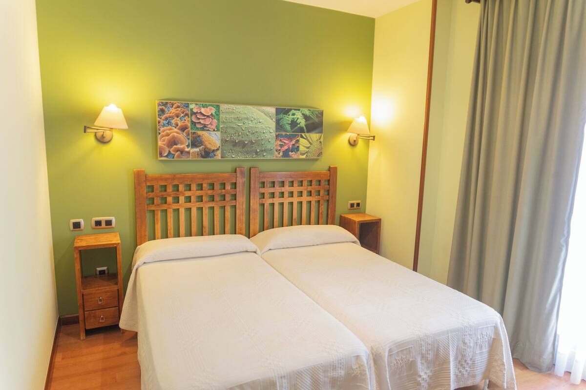 Un lit double avec une tête de lit en bois, deux tables de chevet, une lampe fixée au mur et une œuvre d’art botanique encadrée.