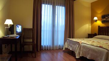 Habitación de hotel con escritorio, computadora, silla, dos camas con cubrecamas florales, una ventana con cortinas y una lámpara fijada a la pared.