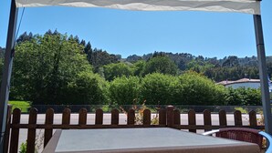 Un área de estar al aire libre cubierta con mesa y sillas, con vistas a una colina con árboles y un edificio.