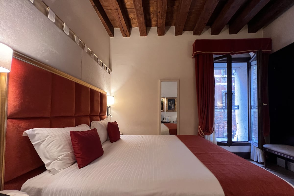 A hotel room with a large bed, a wooden ceiling, a window with red curtains, and a view of the outside.