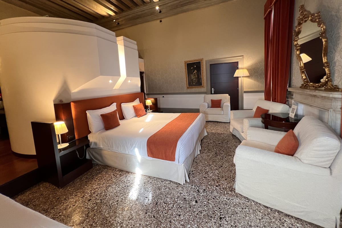 A hotel room with a large bed, two armchairs, a small table, and a fireplace.