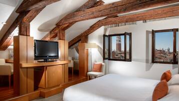 Habitación de hotel con techo de madera, cama con ropa blanca, televisor sobre un mueble de madera, una silla e imágenes enmarcadas en la pared.