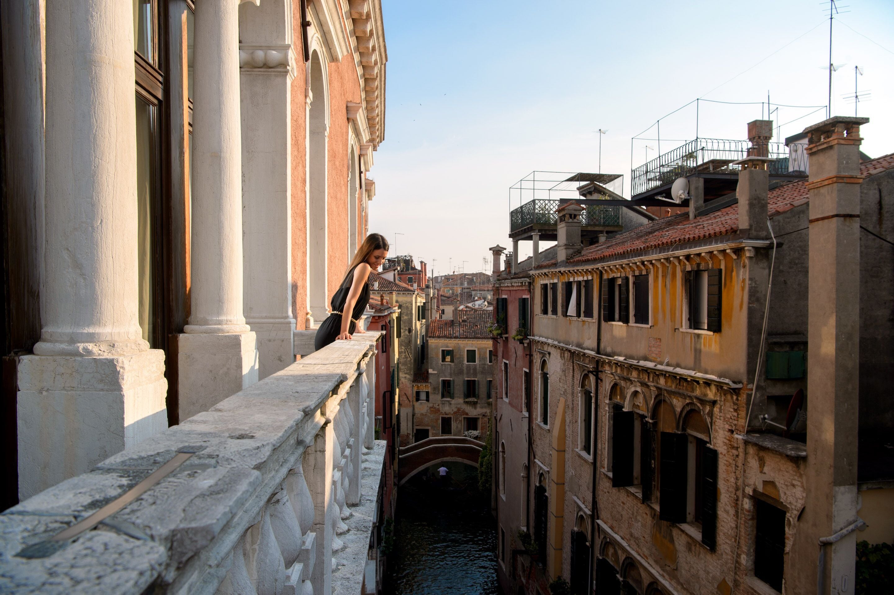 A narrow canal with buildings on either side, a bridge in the distance, and a person leaning on a railing.