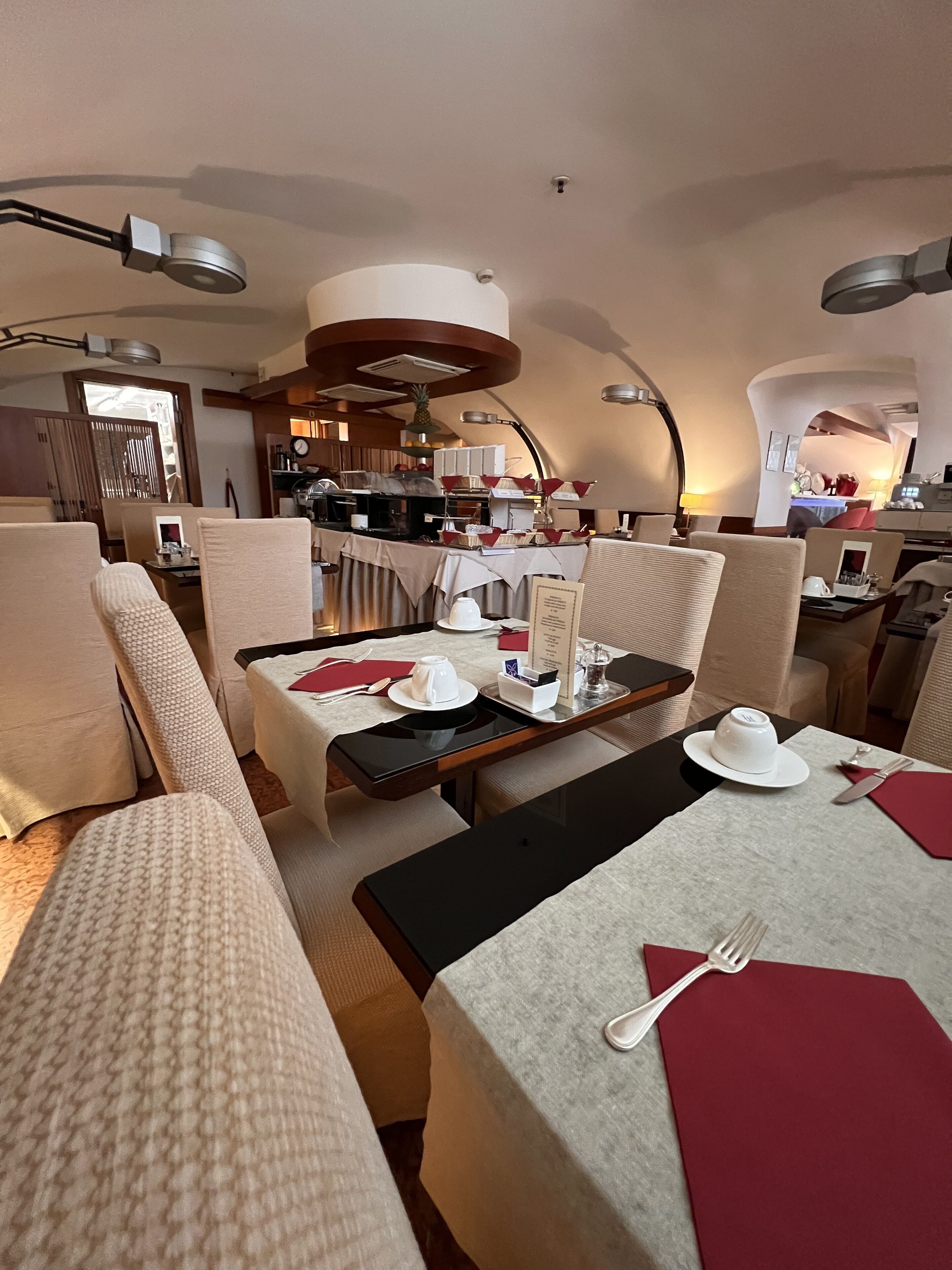A hotel dining area with tables set for two, featuring a modern design with neutral tones and a central island.