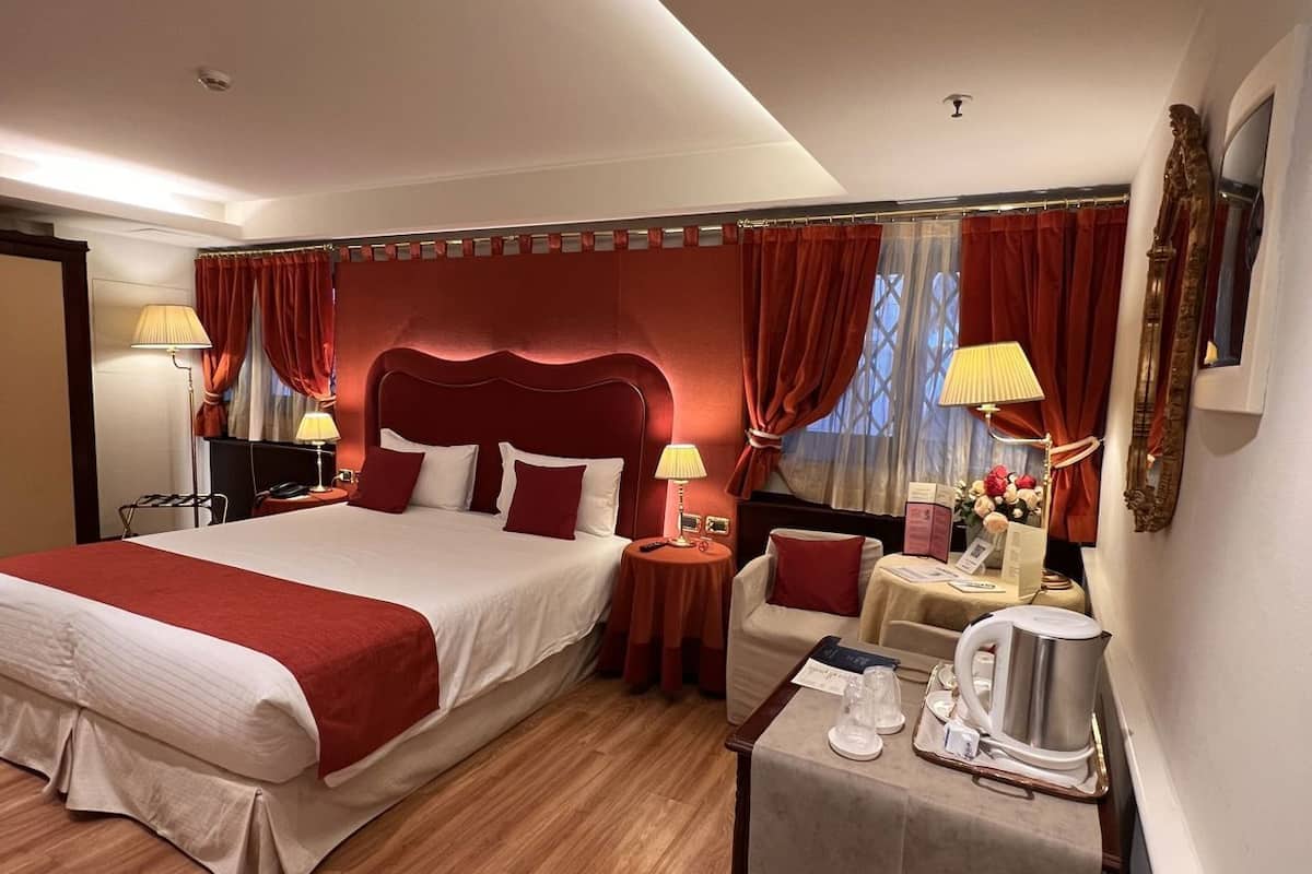 A hotel room with a large bed, two bedside tables, a small table with a kettle, and a trash bin.