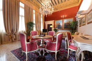Um quarto com uma mesa redonda, cadeiras estofadas em vermelho, um candelabro e uma pintura grande na parede.
