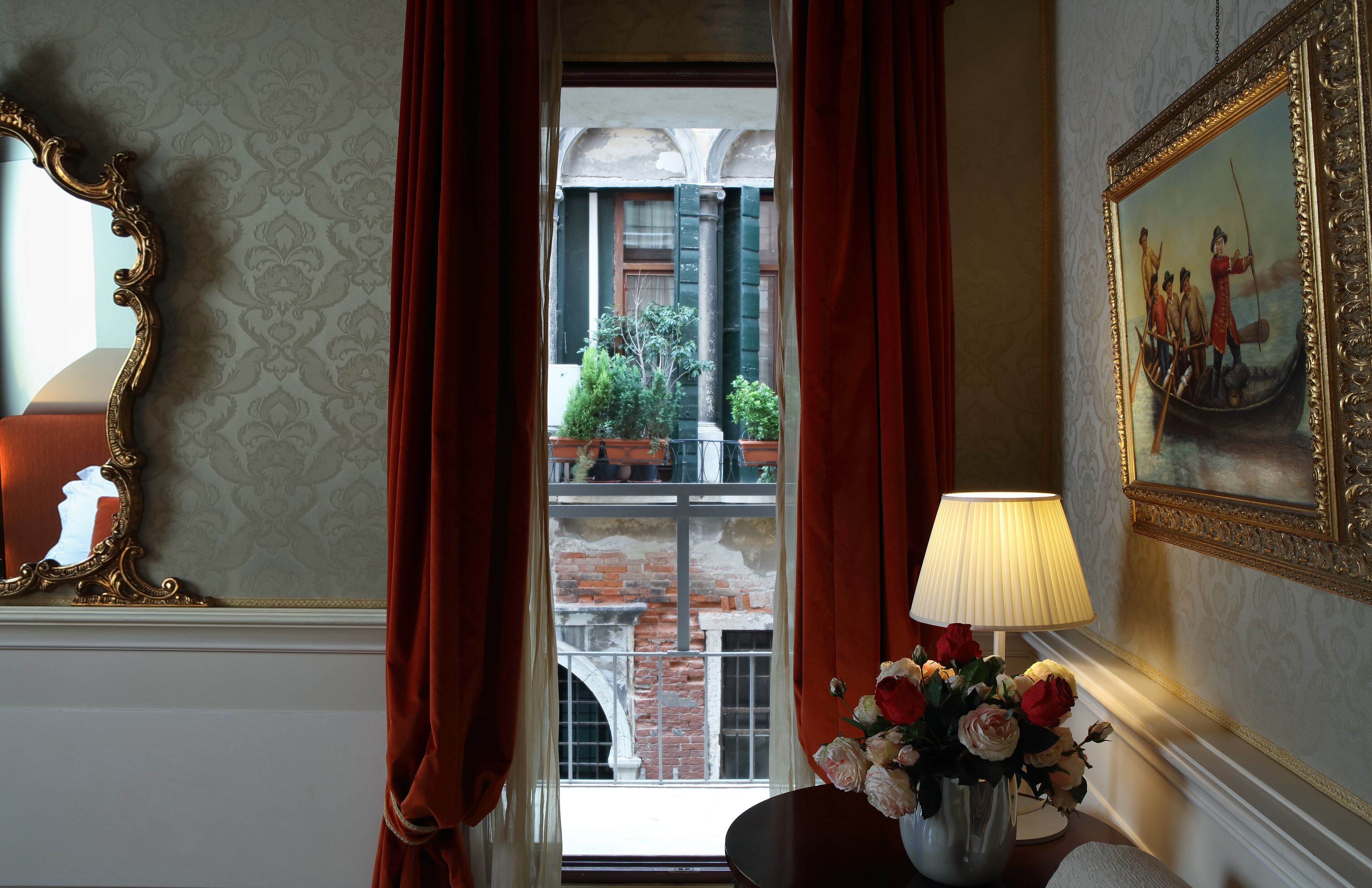 A room with a view of a brick building through a window with red curtains, a table with a lamp and flowers, and a framed painting on the wall.