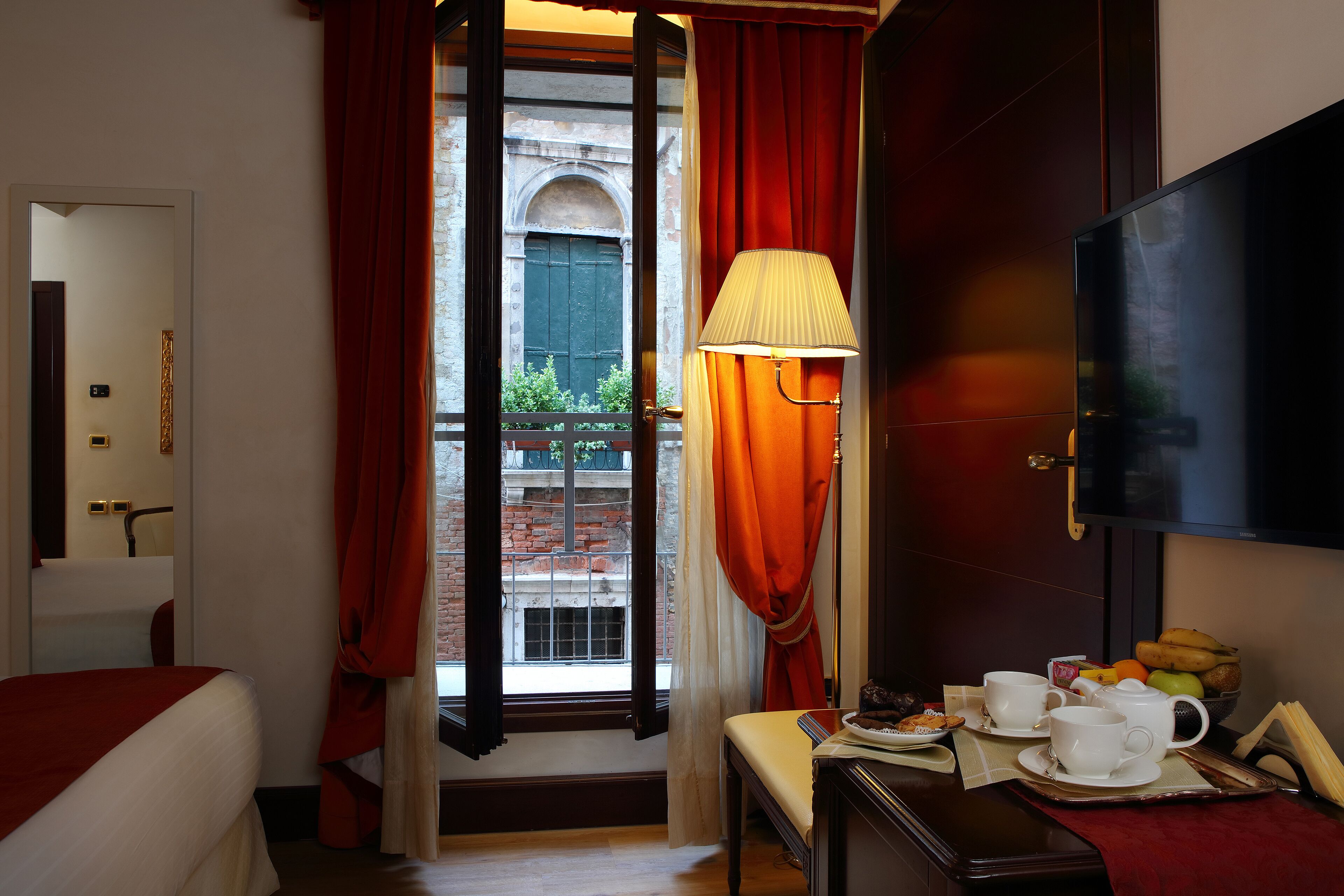 Quarto de hotel com cama, mesa com itens de café da manhã, televisão e janela com cortinas vermelhas.